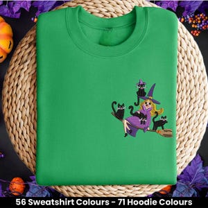May include: Green sweatshirt featuring an embroidered design of a witch riding a broomstick with black cats. The design is in black and purple.