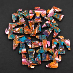 Puede incluir: Una colección de tallas de piedra planas con forma de ave que presentan un vibrante patrón de mosaico de vetas minerales turquesas, naranjas, moradas y blancas. Cada pieza muestra una textura veteada multicolor única con inclusiones de color cobre.