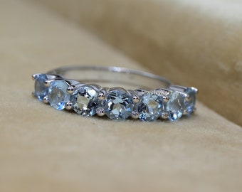 May include: A silver ring featuring seven round, light blue gemstones. The gemstones are set closely together, creating a continuous row across the top of the ring. The ring is displayed on a neutral, beige background.