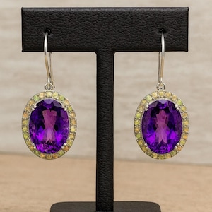 Könnte beinhalten: Ohrringe mit Amethyst-Tropfen und Silberhaken. Jeder Ohrring hat einen großen, ovalen, tiefvioletten Edelstein, umgeben von kleineren, hellgrünen und gelben Steinen. Die Ohrringe werden auf einem schwarzen Ständer mit dem Text "JewelryKart" präsentiert.
