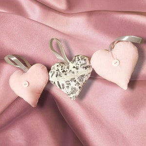 May include: Three heart-shaped decorations with grey ribbon loops. Two are pale pink with a button, and one has a black and white floral pattern with lace trim. They rest on a pink satin background.