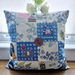 May include: A blue and white patchwork cushion with various embroidered patches, including a pink heart with the text "Girl Gang", a blue elephant with a red balloon, and a brown leather patch with a bee design.