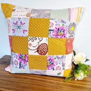 May include: A yellow and brown patchwork cushion with a cartoon pig, butterflies, flowers, and the text "Let's find... acorn" on the front.