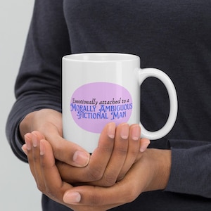 May include: White ceramic mug with a large handle, featuring the text "Emotionally attached to a MORALLY AMBIGUOUS FICTIONAL MAN" in blue and purple on a light purple oval. The mug is held in both hands.