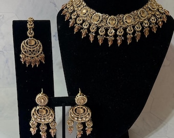 May include: A gold-toned jewelry set, including a necklace and earrings, displayed on black stands. The necklace features a circular design with dangling accents. The earrings have a similar design, with multiple tiers and dangling beads. The set is against a white and gray background.
