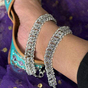May include: A foot adorned with a turquoise shoe and a silver anklet. The shoe has detailed embroidery. The anklet is made of clear, oval-shaped stones. The background is a purple fabric.