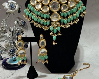 May include: A gold and silver necklace set with green beads and clear stones. The necklace is on a black stand. The earrings and a maang tikka are also included.