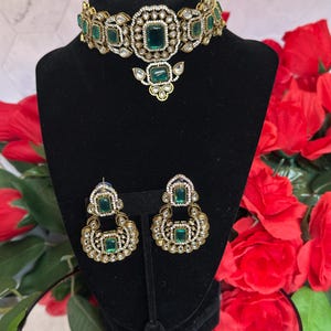 Puede incluir: Un conjunto de collar y pendientes dorados con piedras preciosas de color verde esmeralda y transparentes. El collar es de estilo gargantilla con un colgante central. Los pendientes son de estilo colgante. Las joyas se exhiben en un maniquí negro.