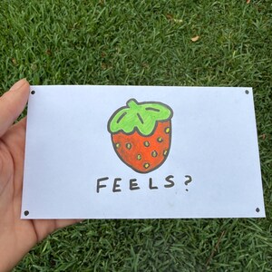 May include: A white rectangular card with a hand-drawn strawberry illustration. The strawberry is red with green leaves and small yellow seeds. The word "FEELS?" is written below the strawberry in black letters.