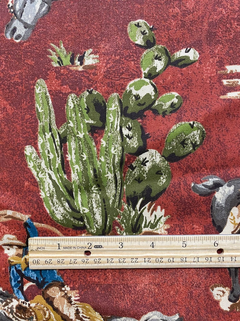 May include: Red fabric with a repeating pattern of green cactus plants and cowboys riding horses. The fabric is measured with a ruler that reads "MADE IN CHINA".