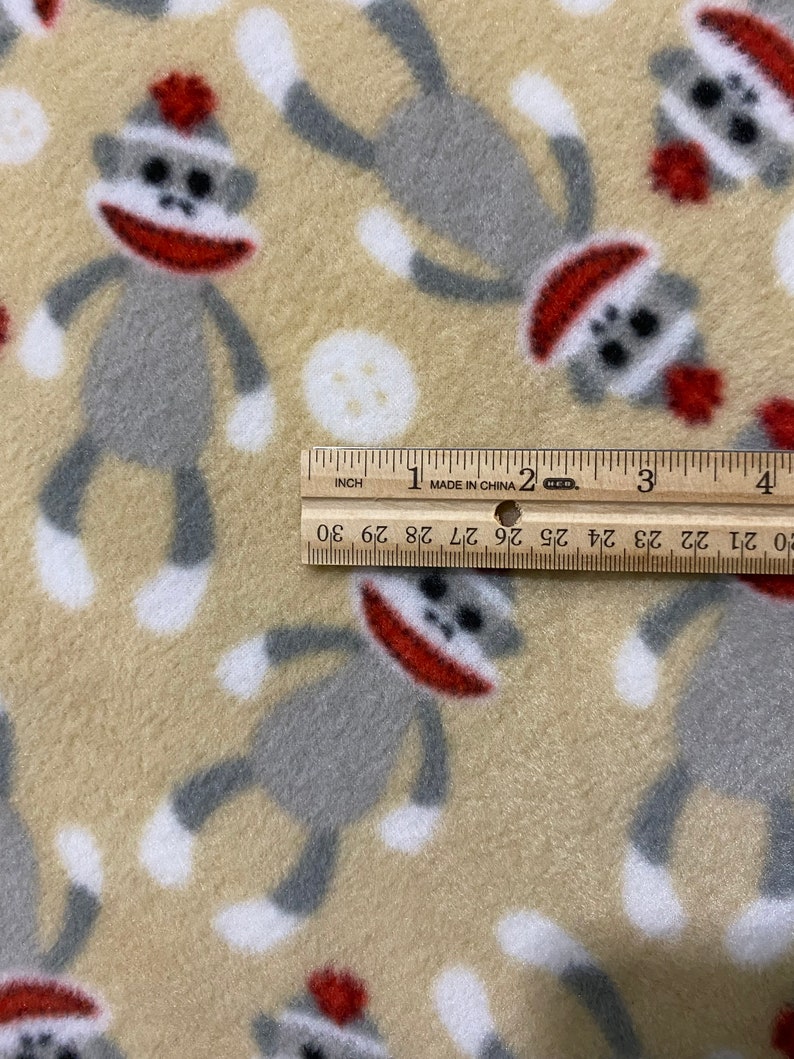 May include: Beige fleece fabric with a repeating pattern of gray sock monkeys wearing red hats. A ruler is placed on the fabric, showing the measurement of 30 inches.
