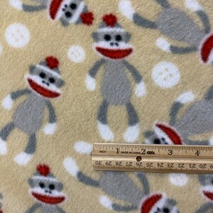 May include: Beige fleece fabric with a repeating pattern of gray sock monkeys wearing red hats. The monkeys are standing with their arms outstretched and legs spread apart. There are also white polka dots scattered throughout the fabric.