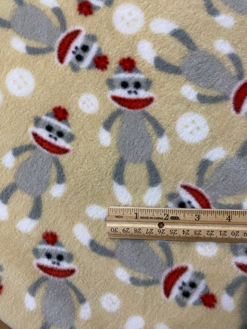May include: Beige fleece fabric with a repeating pattern of gray sock monkeys wearing red hats. The monkeys are standing with their arms outstretched and legs spread apart. There are also white polka dots scattered throughout the fabric.