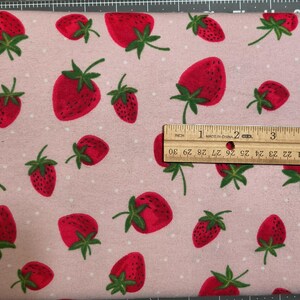 May include: Pink fabric with a repeating pattern of red strawberries and white polka dots. A ruler shows the fabric is approximately 3 inches wide. The strawberries have green leaves.