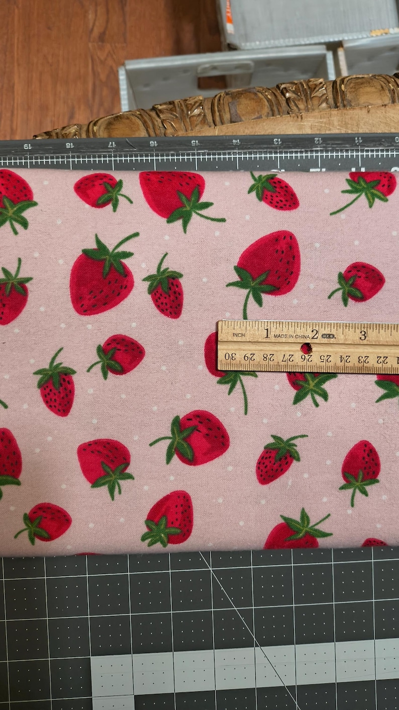 May include: Pink fabric with a repeating pattern of red strawberries and white polka dots. A ruler shows the fabric is approximately 3 inches wide. The strawberries have green leaves.