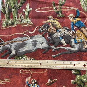 May include: A red fabric with a repeating pattern of cowboys riding horses and lassoing cattle. The fabric features a brown, green, and white color scheme. The fabric is printed with the text "AN AUTHENTIC W".