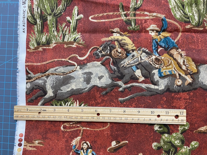 May include: A red fabric with a repeating pattern of cowboys riding horses and lassoing cattle. The fabric features a brown, green, and white color scheme. The fabric is printed with the text "AN AUTHENTIC W".