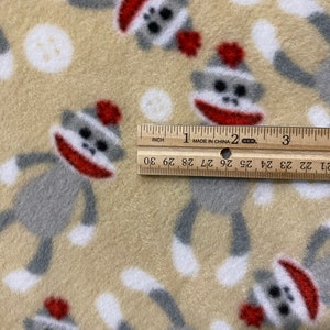 May include: Beige fleece fabric with a repeating pattern of gray and white monkeys wearing red hats. The monkeys are in a playful pose with their arms and legs outstretched. The fabric is measured with a ruler, showing 3 inches.