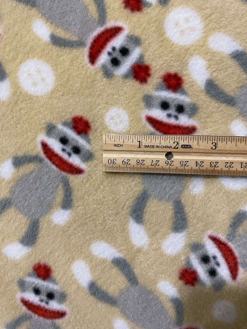 May include: Beige fleece fabric with a repeating pattern of gray and white monkeys wearing red hats. The monkeys are in a playful pose with their arms and legs outstretched. The fabric is measured with a ruler, showing 3 inches.