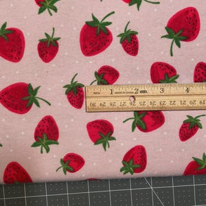 May include: Pink fabric with a repeating pattern of red strawberries with green leaves and white polka dots. A wooden ruler shows inches. The fabric is on a black and gray cutting mat.