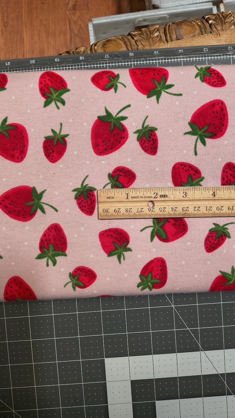 May include: Pink fabric with a repeating pattern of red strawberries with green leaves and white polka dots. A wooden ruler shows inches. The fabric is on a black and gray cutting mat.