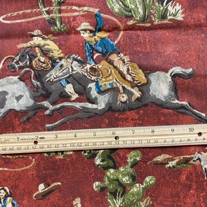 May include: A red fabric with a repeating pattern of cowboys riding horses. The cowboys are wearing hats and boots. The horses are brown and white. The fabric has a rustic, vintage look.