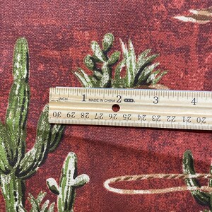 May include: Close-up of a red fabric with a repeating pattern of cacti and cowboys. A ruler is placed on the fabric to show the scale. The ruler reads "MADE IN CHINA" and has markings in inches.