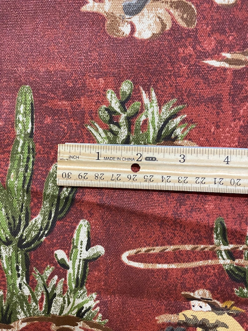 May include: Close-up of a red fabric with a repeating pattern of cacti and cowboys. A ruler is placed on the fabric to show the scale. The ruler reads "MADE IN CHINA" and has markings in inches.