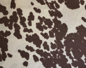 May include: A fabric featuring a repeating cow print pattern with irregular dark brown spots scattered across a light cream-colored background. The material displays a consistent texture and a high-contrast design that mimics the natural hide of a cow.