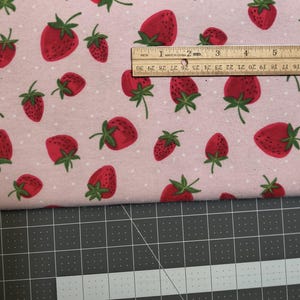 May include: Pink fabric with a repeating pattern of red strawberries with green stems and leaves. The fabric is measured with a wooden ruler, showing inches. The strawberries are of varying sizes and are scattered across the fabric.
