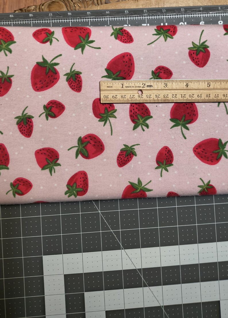 May include: Pink fabric with a repeating pattern of red strawberries with green stems and leaves. The fabric is measured with a wooden ruler, showing inches. The strawberries are of varying sizes and are scattered across the fabric.