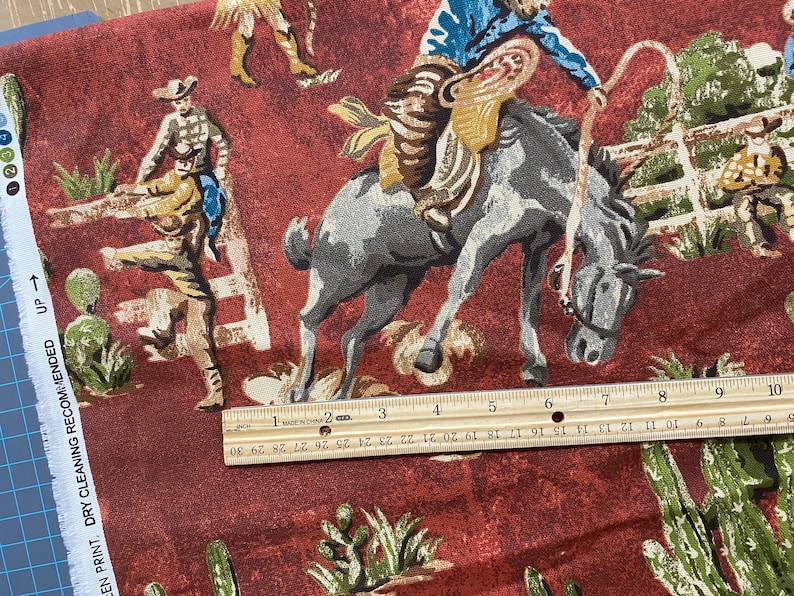 May include: Red fabric with a repeating pattern of cowboys riding horses and cacti. The fabric is 10 inches wide.