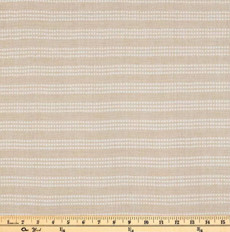 Op de afbeelding: Een close-up van een beige stof met witte strepen en kleine witte stippen. De stof wordt gemeten met een liniaal in inches.