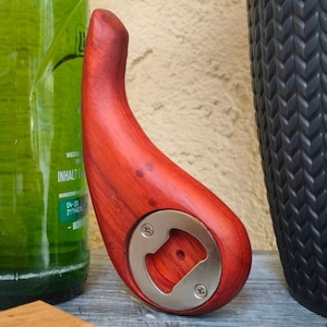 Puede incluir: Un abridor de botellas de madera roja con un abridor de tapas de metal plateado. El abridor tiene forma de pera y tiene una veta de madera natural.