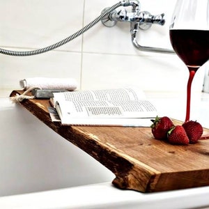 Puede incluir: Una bandeja de baño de madera con un borde natural, que incluye un libro, una copa de vino tinto y fresas. La bandeja está colocada sobre una bañera blanca.