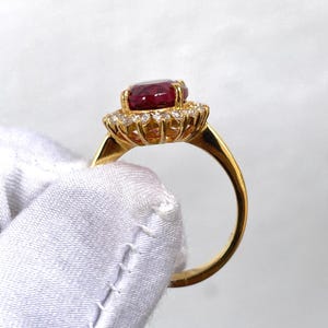 Puede incluir: Un anillo de oro con una gran gema roja oscura de corte ovalado, rodeada de gemas m&aacute;s peque&ntilde;as y transparentes. La banda del anillo es de oro liso. El anillo se muestra sobre un fondo blanco.