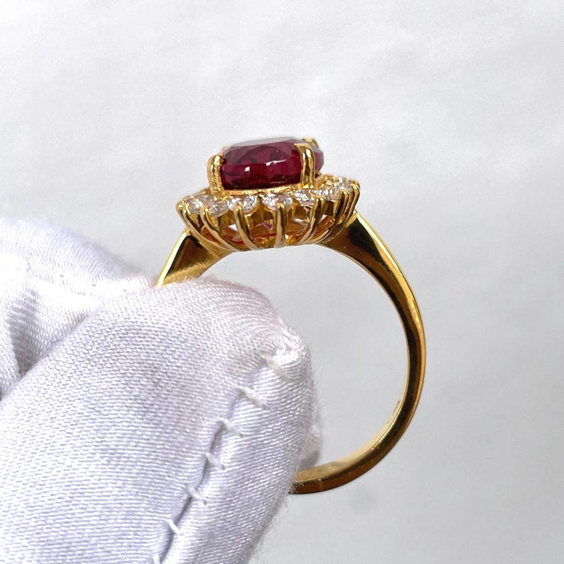 Puede incluir: Un anillo de oro con una gran gema roja oscura de corte ovalado, rodeada de gemas m&aacute;s peque&ntilde;as y transparentes. La banda del anillo es de oro liso. El anillo se muestra sobre un fondo blanco.