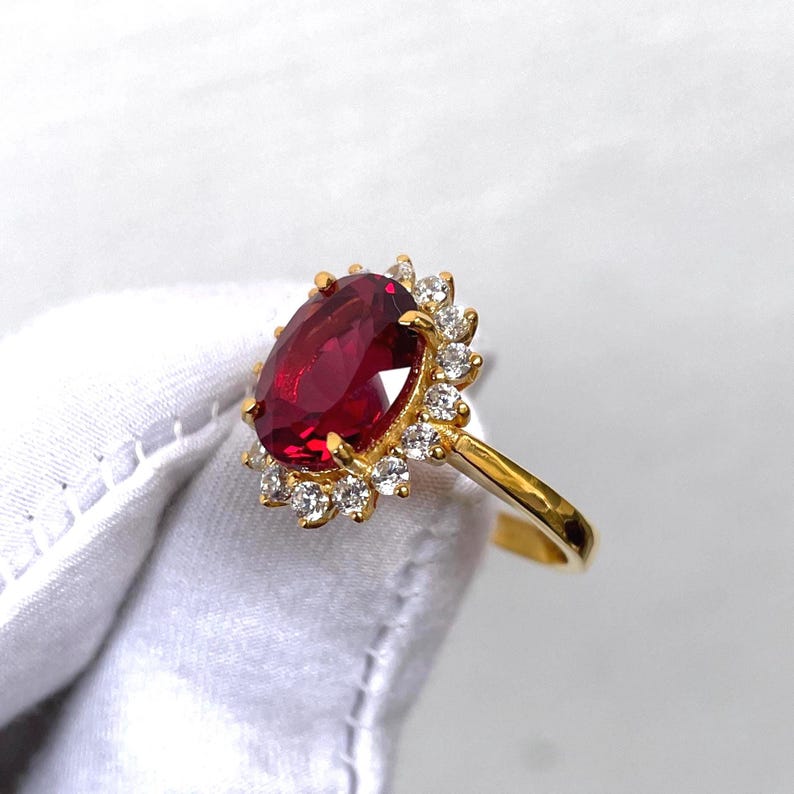 Puede incluir: Un anillo de oro con una gran gema roja oscura de corte ovalado, rodeada de peque&ntilde;as gemas claras. La banda del anillo es de oro liso. Las gemas est&aacute;n dispuestas en un halo, creando un efecto brillante. El anillo se muestra sobre un fondo blanco.