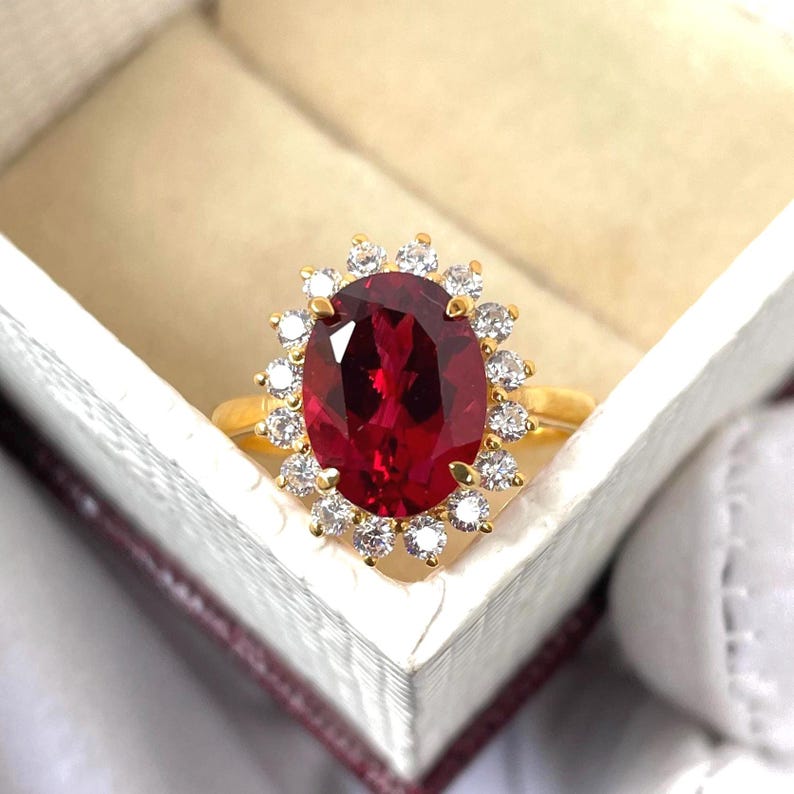 Puede incluir: Un anillo de oro con una gran gema roja oscura de corte ovalado, rodeada de piedras m&aacute;s peque&ntilde;as y claras. El anillo se presenta en una caja de joyer&iacute;a blanca con interior granate, resaltando su brillo.