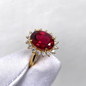 Puede incluir: Un anillo de oro con una gran piedra preciosa roja oscura de corte ovalado, rodeada por un halo de piedras preciosas m&aacute;s peque&ntilde;as y claras. La banda del anillo es de oro liso. Las piedras preciosas est&aacute;n engastadas sobre un fondo blanco.