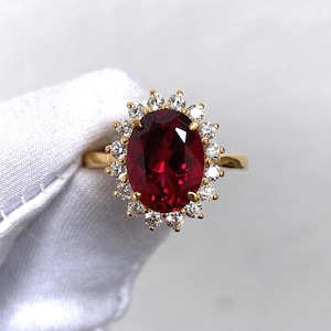 May include: A gold ring featuring a large, oval-cut, deep red gemstone surrounded by a halo of smaller, clear gemstones. The ring's band is a simple, polished gold. The gemstones are set against a white background.