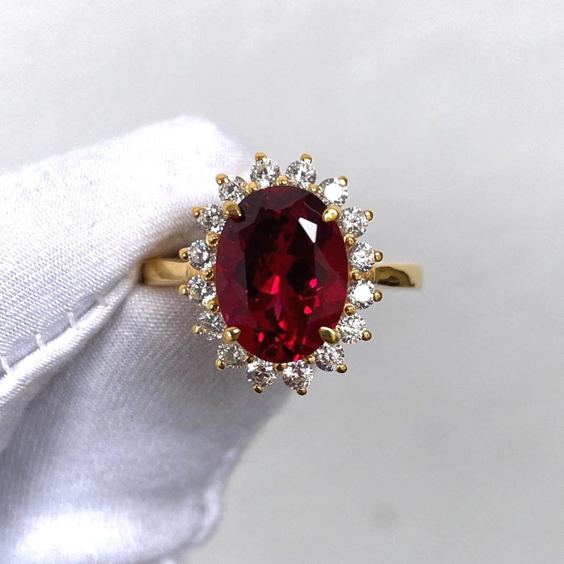 Puede incluir: Un anillo de oro con una gran gema roja oscura de corte ovalado, rodeada por un halo de gemas m&aacute;s peque&ntilde;as y claras. La banda del anillo es de oro pulido. Las gemas est&aacute;n engastadas sobre un fondo blanco.