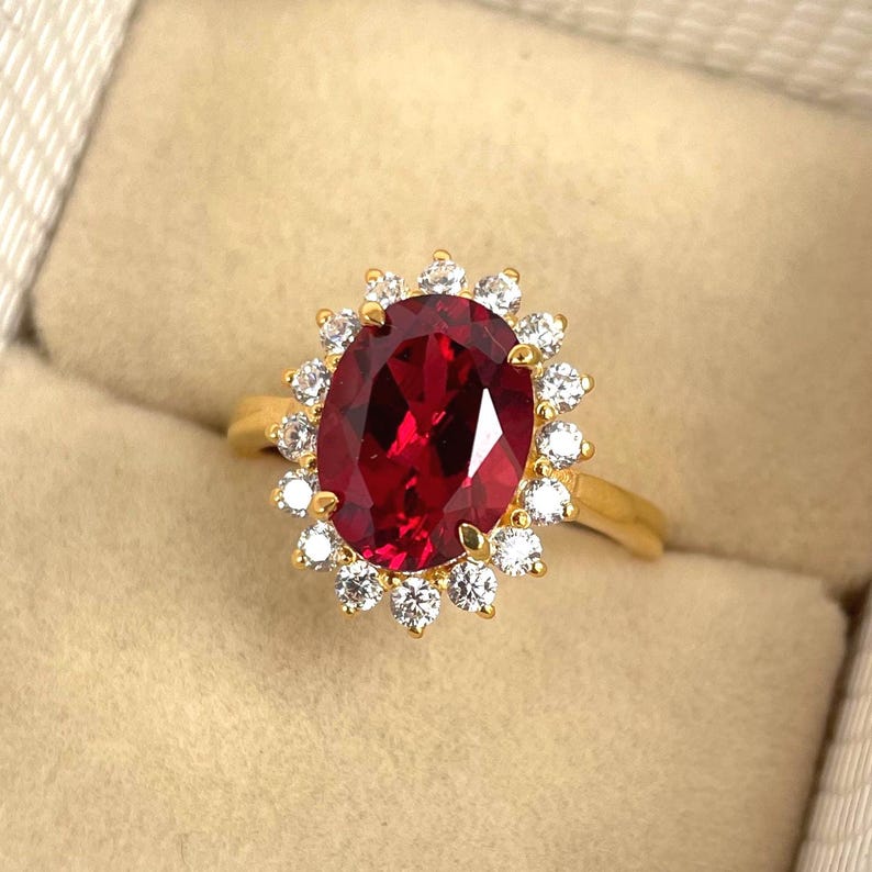 Puede incluir: Un anillo de oro con una gran gema roja oscura de forma ovalada, rodeada de gemas claras m&aacute;s peque&ntilde;as. El anillo tiene una banda de oro lisa. El engaste resalta el brillo de la piedra central. Joya.