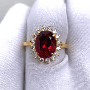 Puede incluir: Un anillo de oro con una gran gema roja oscura de corte ovalado, rodeada por un halo de gemas m&aacute;s peque&ntilde;as y claras. La banda del anillo es de oro liso. El anillo se muestra sobre un fondo blanco.
