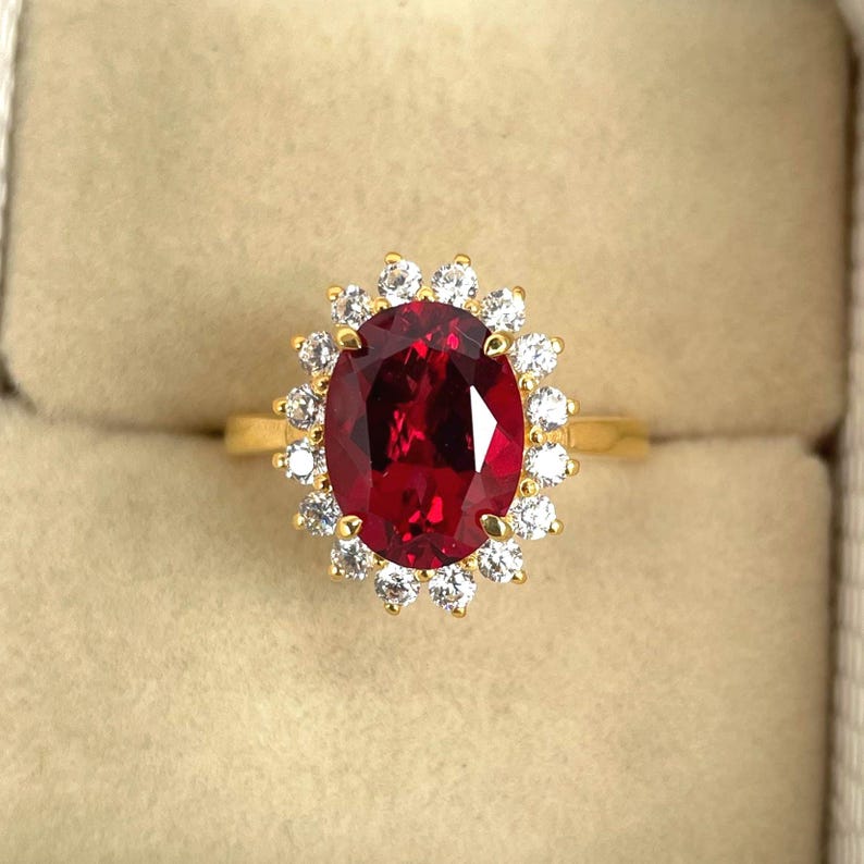 Puede incluir: Un anillo de oro con una gran gema roja oscura de corte ovalado, rodeada por un halo de gemas claras m&aacute;s peque&ntilde;as y redondas. El anillo tiene una banda de oro pulido. El dise&ntilde;o resalta el color vibrante de la piedra central.