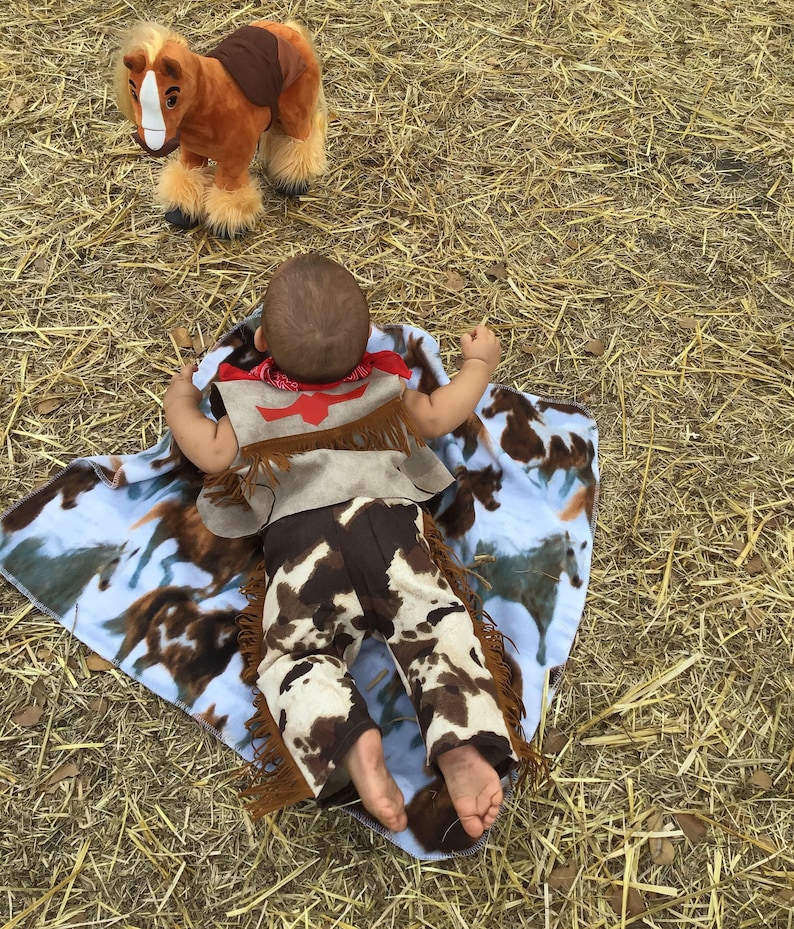 May include: A baby wearing a brown and white cowboy outfit with a red bandana and fringe lies on a blue and white blanket with a brown horse toy in the background.