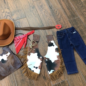 May include: A child's cowboy costume set, including a brown hat, red bandana, brown vest, cow-print chaps with fringe, a brown belt with a bull buckle, and blue jeans. The costume is designed for dress-up and imaginative play.