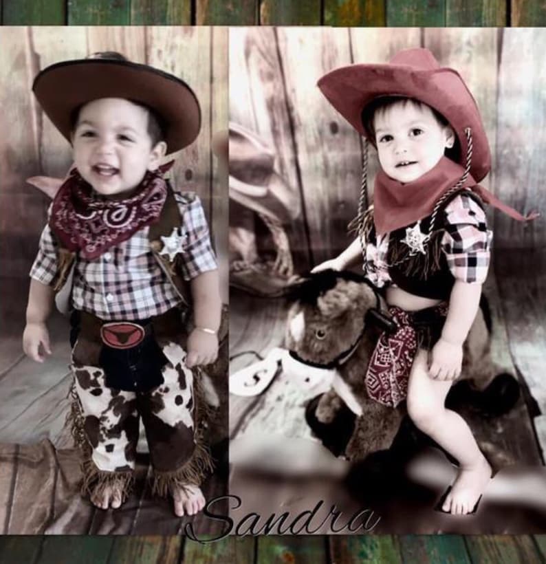 May include: Two toddlers dressed in cowboy costumes. One is wearing a plaid shirt, brown pants with fringe, a red bandana, and a brown cowboy hat. The other is wearing a plaid shirt, a red bandana, and a red cowboy hat. They are both smiling.