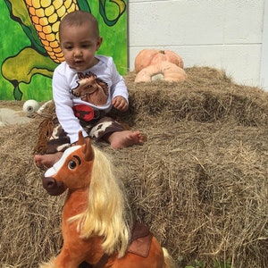 May include: A young child dressed in a cowboy costume sits on a plush brown horse toy. The child is wearing a white long-sleeved shirt with a brown horse graphic and brown and white cowboy pants. The child is sitting on a pile of hay with pumpkins in the background.