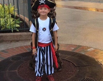 May include: A young boy dressed as a pirate wearing a black and white striped vest, black and white striped pants, a red sash, a white t-shirt with a black skull and crossbones design, a black pirate hat with a gold skull and crossbones design, and a red bandana.